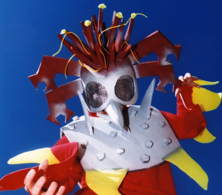 A child in a spiky, red and yellow costume, with silver chestplate and mask with huge eyes and long pointy nose.